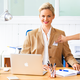 Een vrouw met blond haar zit aan een bureau, glimlacht en draagt een blauwe blazer. Een laptop en schrijfgerei zijn zichtbaar.