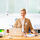 Een vrouw met blond haar zit achter een bureau, glimlacht en draagt een blauwe blazer. Voor haar staat een laptop en schrijfmaterialen.