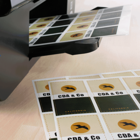 Ein Drucker gibt Etiketten mit dem Text "CDA & CO" und "CALIFORNIA" aus, die auf einem Tisch liegen.
