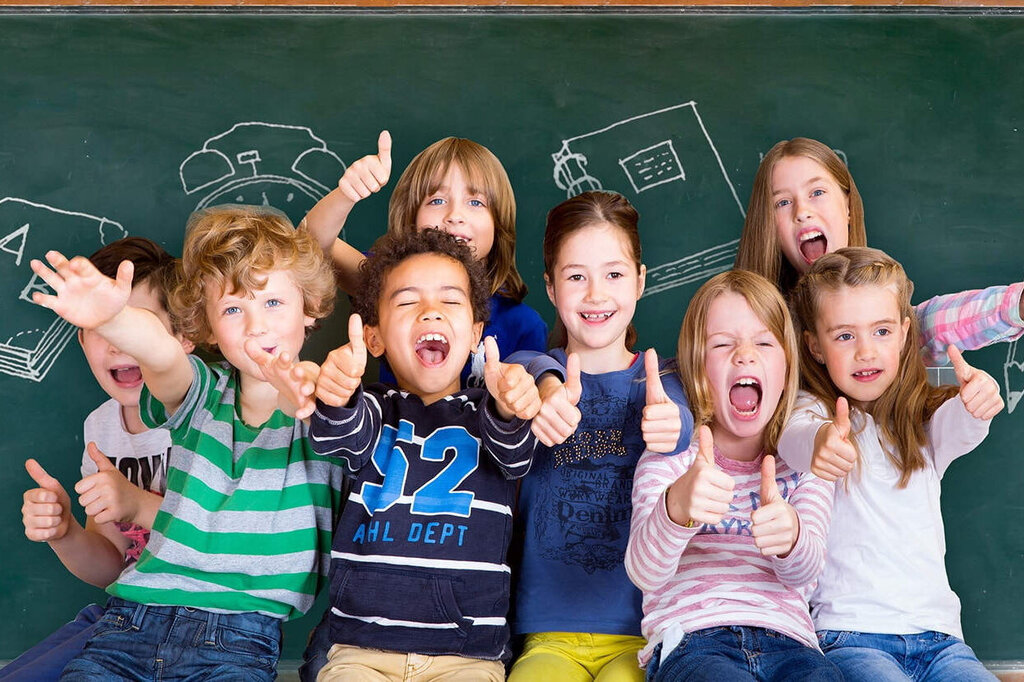 Een groep kinderen staat voor een bord, glimlacht en poseert vrolijk. Tekeningen van schoolmaterialen zijn op de achtergrond zichtbaar.
