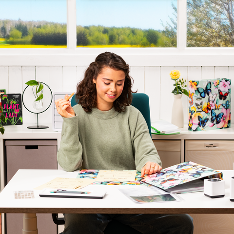 Een jonge vrouw zit aan een tafel, omringd door notities en een laptop, terwijl ze met één hand een gebaar maakt. Op de achtergrond zijn ramen met uitzicht op een groene landschap.