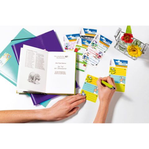 Eine Hand schreibt in ein Notizbuch, umgeben von bunten Aufklebern und einem Blumenarrangement. Ein Buch mit Illustrationen ist geöffnet.