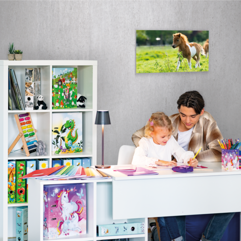 Ein Mädchen sitzt an einem Schreibtisch, schreibt in ein Heft und hat einen bunten Stiftkasten vor sich. Im Hintergrund sind Regale mit Spielzeug und Kunstmaterialien.