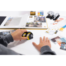 Eine Hand hält ein gelbes und schwarzes Gerät über ein Blatt Papier, umgeben von verschiedenen Dokumenten, Fotos und Bürobedarf.