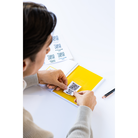 Eine Person klebt einen QR-Code auf eine gelbe Seite eines Notizbuchs, während sie einen Stift in der Hand hält.