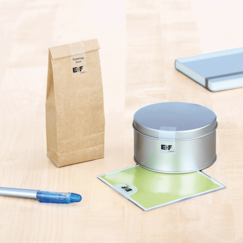 Ein silbernes Metallbehälter, ein brauner Papierbeutel, ein grünes Blatt und ein blauer Stift auf einem Holztisch.