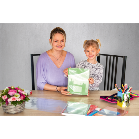 Eine Frau und ein Mädchen sitzen an einem Tisch und halten ein grünes Heft. Auf dem Tisch liegen bunte Stifte und ein Blumenstrauß.