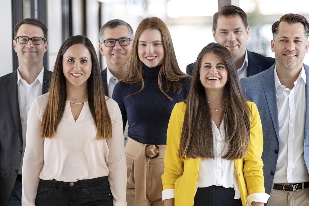 Een groep van zeven personen staat in een gang, glimlachend en in formele kleding, sommigen dragen blouses en pakken.