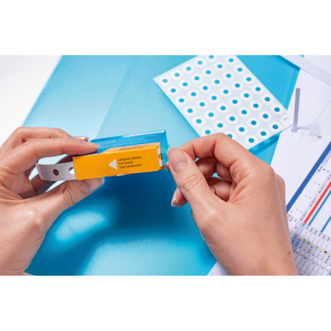 Eine Person hält eine Verpackung mit einem gelben Aufkleber, während sie auf einen blauen Hintergrund und ein Blatt mit Löchern schaut.