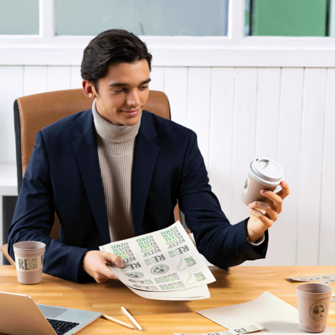 Ein junger Mann in einem Anzug sitzt an einem Tisch, hält einen Kaffeebecher und betrachtet Papiere mit grünen Druckmotiven.