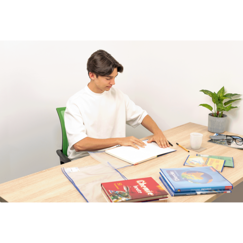 Ein junger Mann sitzt an einem Tisch, blättert in einem Notizbuch und hat mehrere Bücher sowie eine Pflanze und eine Tasse auf dem Tisch.