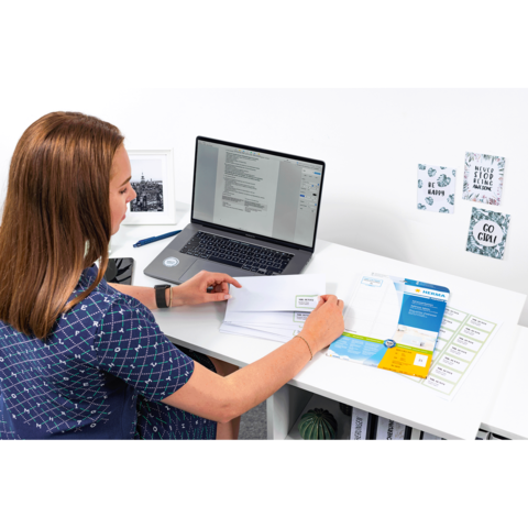 Eine Person sitzt an einem Schreibtisch und bearbeitet Dokumente, während ein Laptop mit geöffnetem Textdokument vor ihr steht.