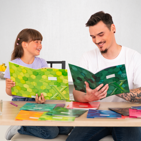 Ein Erwachsener und ein Kind sitzen an einem Tisch und betrachten farbige Hefte. Der Erwachsene zeigt auf ein Heft, während das Kind lächelt.