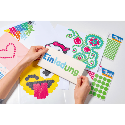 Hände halten eine Einladungskarte mit dem Wort "Einladung" in bunten Buchstaben, umgeben von Bastelmaterialien und Aufklebern.