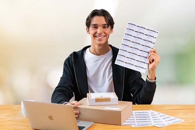 Een jonge man zit aan een tafel met een laptop en houdt een vel met stickers in zijn hand.