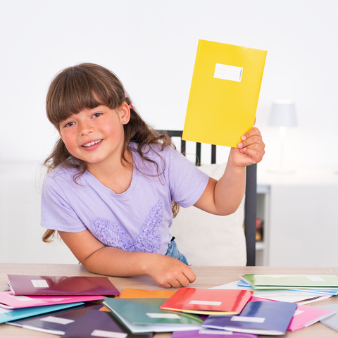 Ein Mädchen hält ein gelbes Heft in der Hand und lächelt, während es auf einem Tisch mit bunten Heften sitzt.