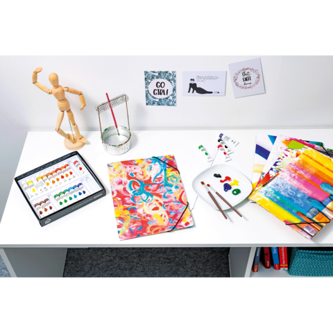 Ein Tisch mit Malutensilien: Aquarellfarben, Pinsel, ein Skizzenblock mit bunten Mustern und inspirierenden Postern im Hintergrund.