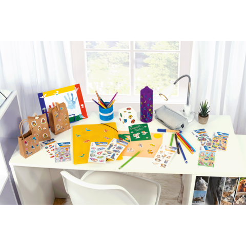 Bastelmaterialien auf einem Tisch, einschließlich Papier, Aufkleber, Stifte und eine Pflanze, mit Fenstern im Hintergrund.