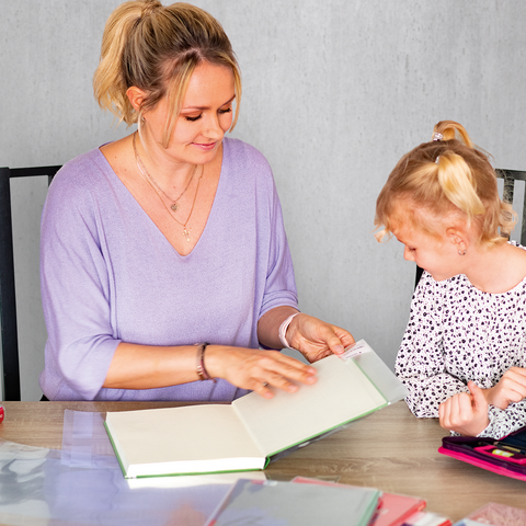 Eine Frau und ein Mädchen sitzen an einem Tisch, schauen in ein Buch und interagieren. Auf dem Tisch liegen verschiedene Schreibmaterialien und Blumen.