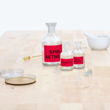 Flasche mit rotem Etikett "Spir. Aether", daneben zwei kleinere Flaschen mit "Barbital" und "Tinct. Tramon", sowie ein Mörser und Petrischalen.