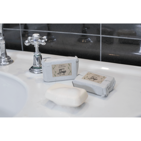 Zwei Seifenstücke, eines verpackt und eines unverpackt, liegen auf einem Waschbecken neben einem Wasserhahn.