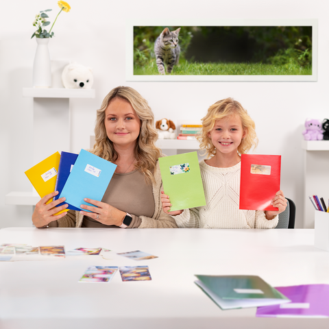 Een vrouw en een meisje houden kleurrijke mappen vast terwijl ze aan een tafel met foto's zitten. Op de achtergrond is een kat te zien.