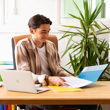 Ein junger Mann sitzt an einem Schreibtisch, arbeitet mit einem Laptop und blättert durch Dokumente in einer Ordner. Pflanzen und bunte Aktenordner sind im Hintergrund sichtbar.
